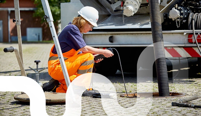 Eine Mitarbeiterin mit Schutzhelm schaut auf das Display ihres Messgerätes, dessen Messfühler in den Abscheider führt. Im Hindergrund steht ein Saug-Druck-Tankfahrzeug.