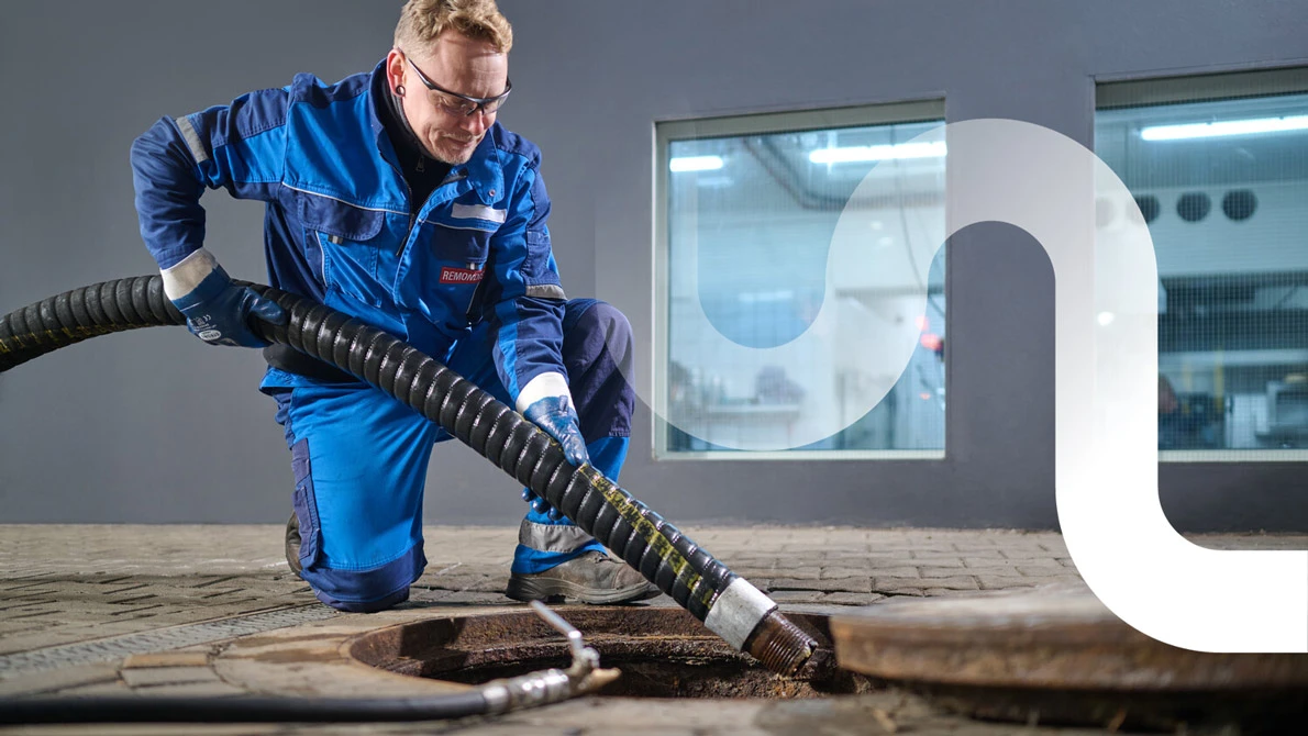 Ein Mitarbeiter von REMONDIS kniet am geöffneten Fettabscheider und führt den Saugschlauch hinein. Im Hintergrund leuchten die Fenster der Restaurantküche