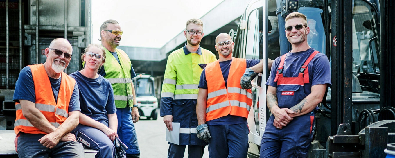 5 Mitarbeiter und 1 Mitarbeiterin von REMONDIS in Arbeitsschutzkleidung sitzen und stehen auf einem Recyclinghof in einer Gruppe zusammen und schauen in die Kamera.