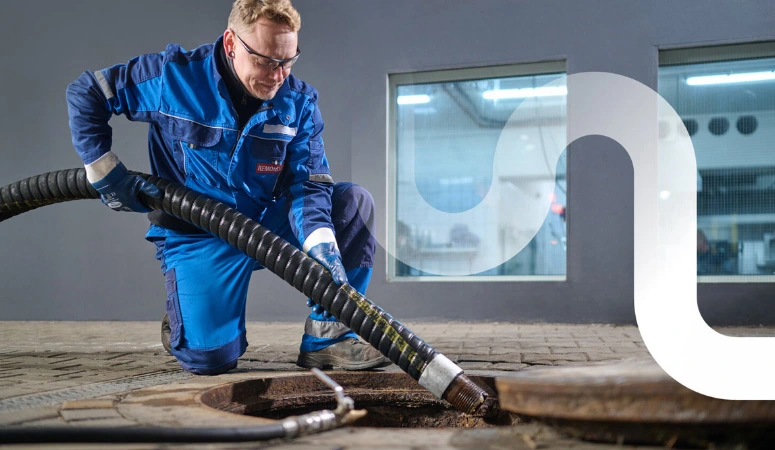Ein Mitarbeiter von REMONDIS kniet am geöffneten Fettabscheider und führt den Saugschlauch hinein. Im Hintergrund leuchten die Fenster der Restaurantküche.
